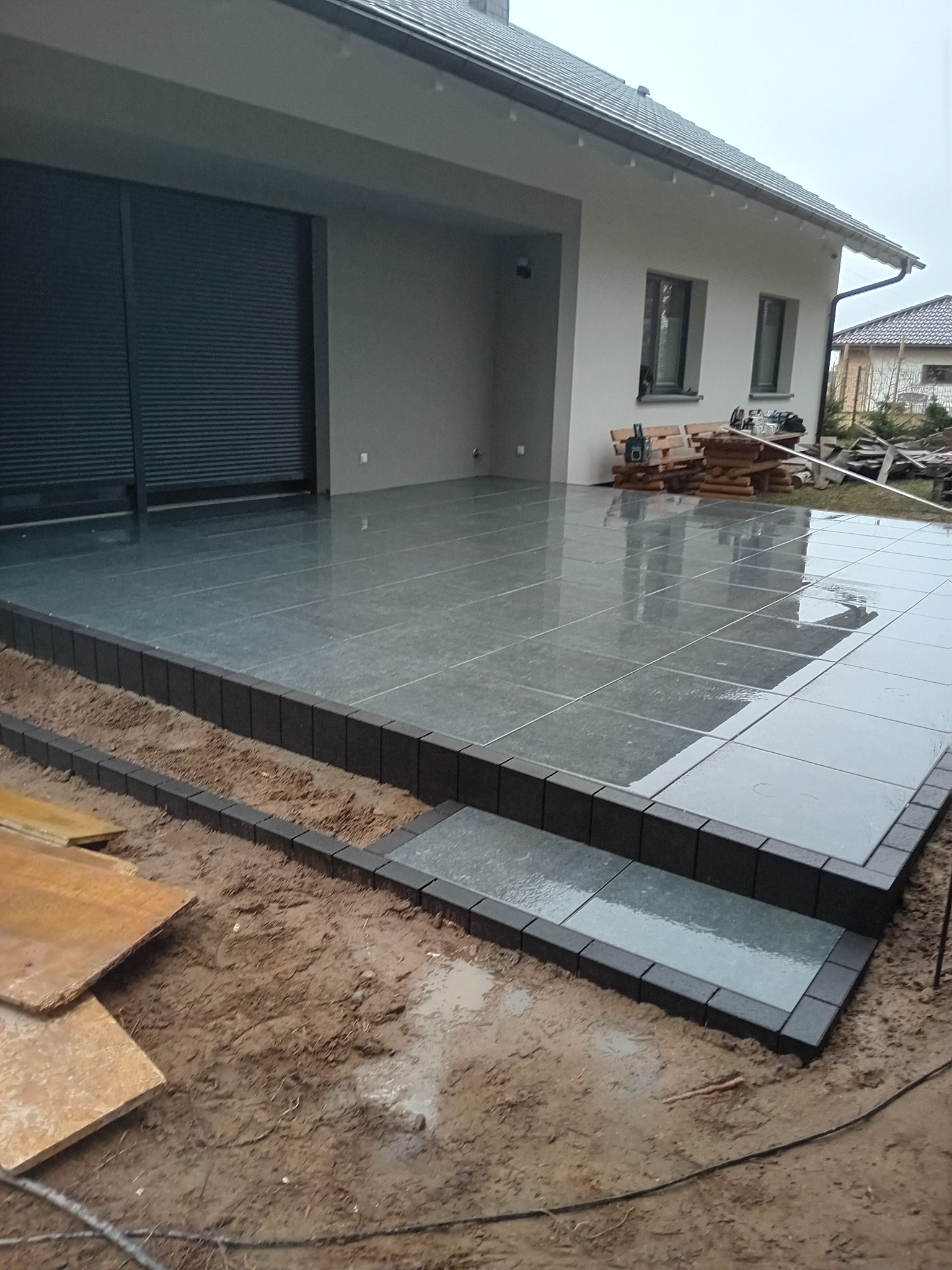 Wykończony taras z szarych płyt z ciemną obwódką, schodki z kostki, widok na nowoczesny dom w pochmurny dzień.