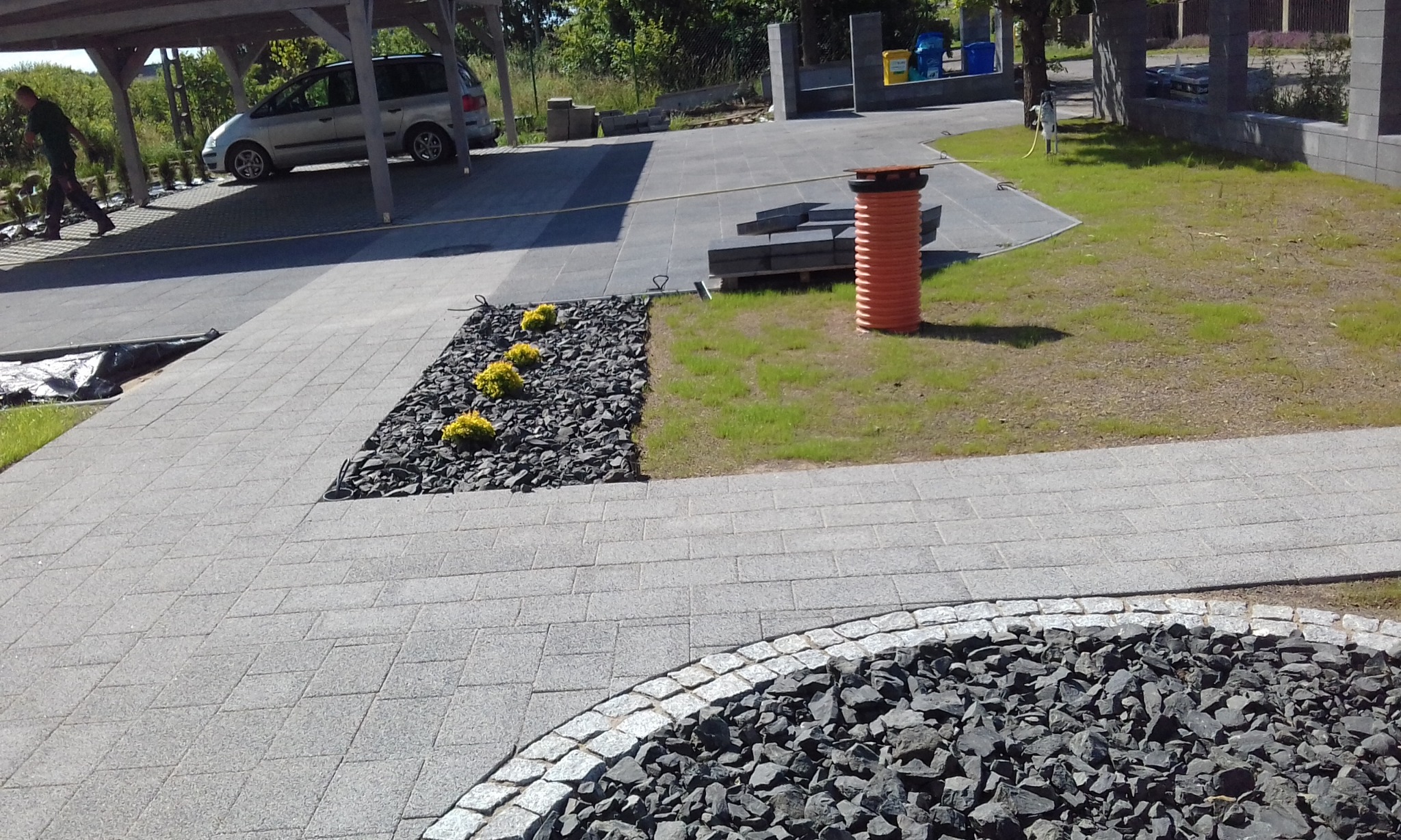 Nowoczesna aranżacja przestrzeni: szara kostka brukowa, czarny grys i żółte krzewy. Widoczna studzienka kanalizacyjna i fragment zaparkowanego samochodu.