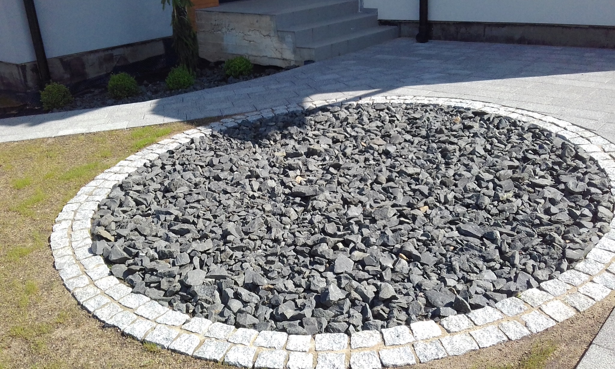 Okrągły klomb z szarego grysu otoczony jasną kostką granitową, widok z góry, w tle fragment domu i chodnik z kostki brukowej.