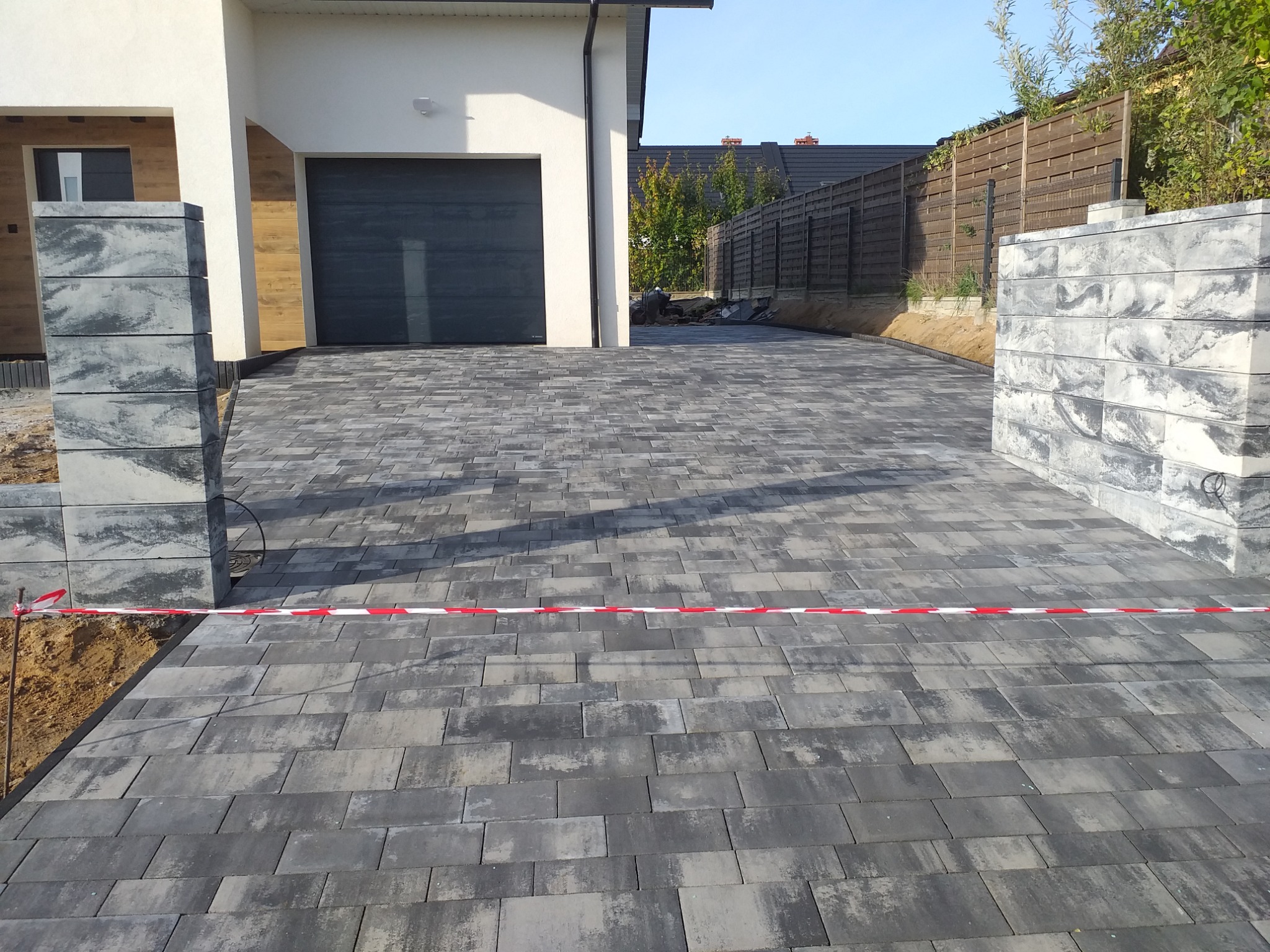 Ułożona szara kostka brukowa prowadząca do garażu nowoczesnego domu. Widoczne słupki z kostki po bokach, taśma ostrzegawcza i fragment drewnianego ogrodzenia w tle.