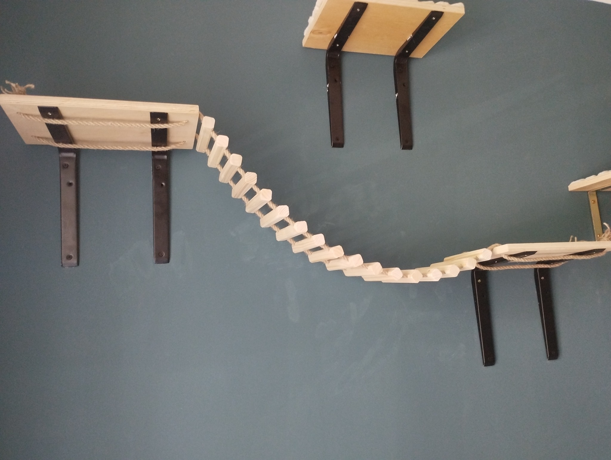 Drewniana konstrukcja półek i mostku linowego, zamontowana na szaro-niebieskiej ścianie. Półki przytwierdzone czarnymi wspornikami. Ujęcie z dołu, widoczne elementy mocujące.