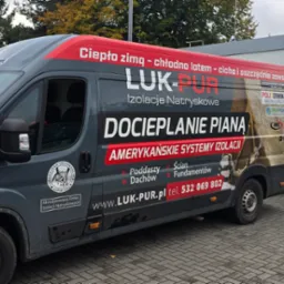 Szary van firmy LUK-PUR z przyczepą i agregatem do natrysku piany, zaparkowany na kostce brukowej przy budynku. Reklama na vanie: 'Docieplanie pianą'.
