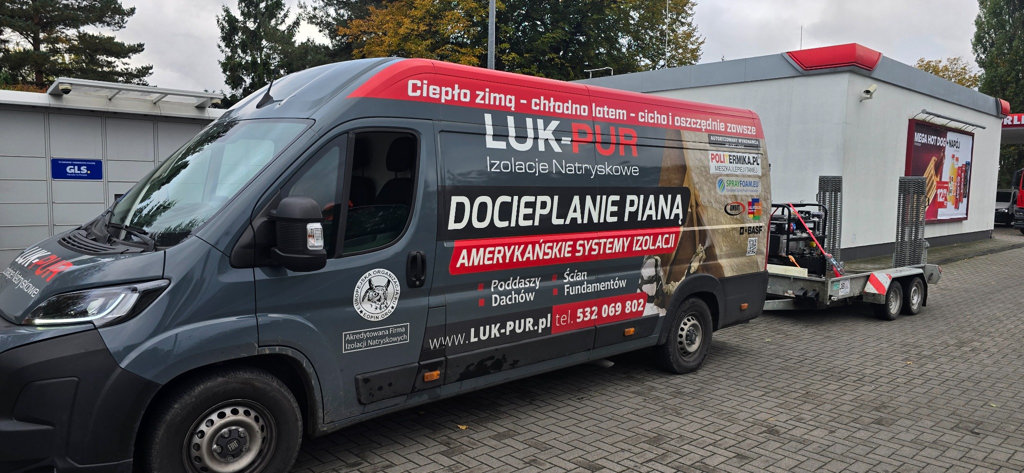 Szary van firmy LUK-PUR z przyczepą i agregatem do natrysku piany, zaparkowany na kostce brukowej przy budynku. Reklama na vanie: 'Docieplanie pianą'.