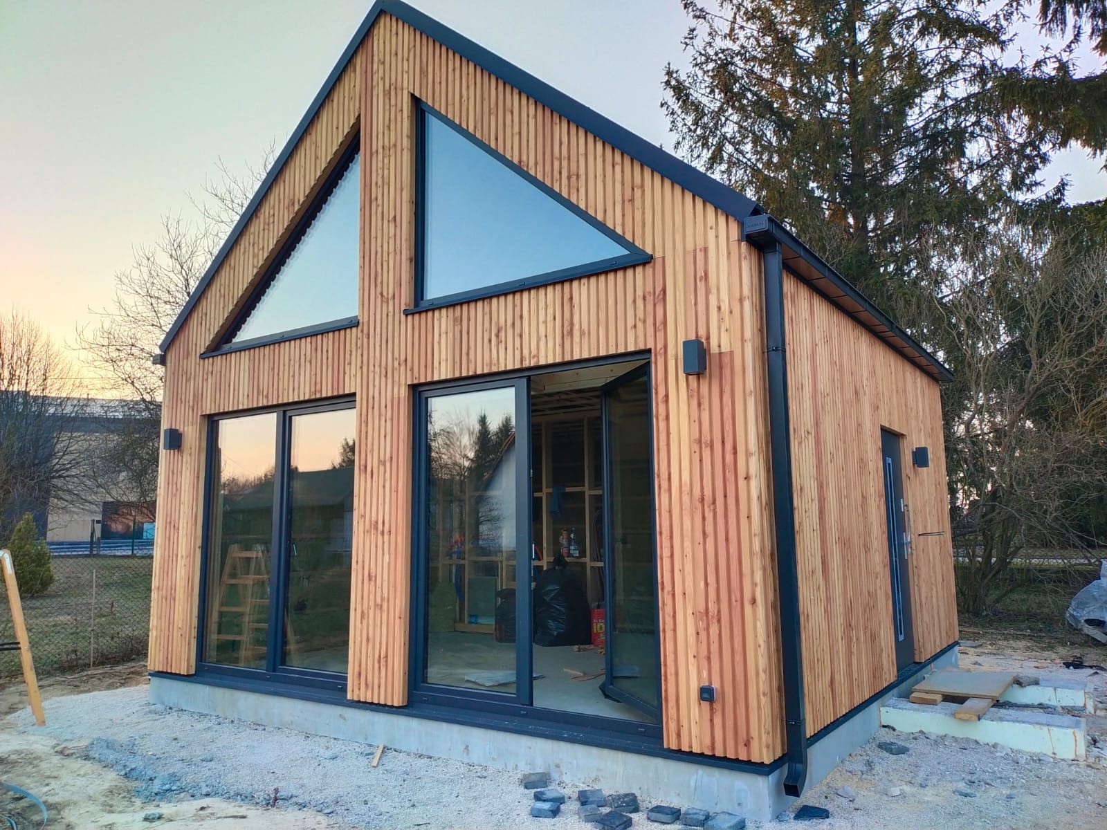 Nowoczesny dom szkieletowy z drewnianą elewacją, dużymi oknami i trójkątnymi przeszkleniami. Widoczne detale wykończenia i otoczenie w trakcie prac.