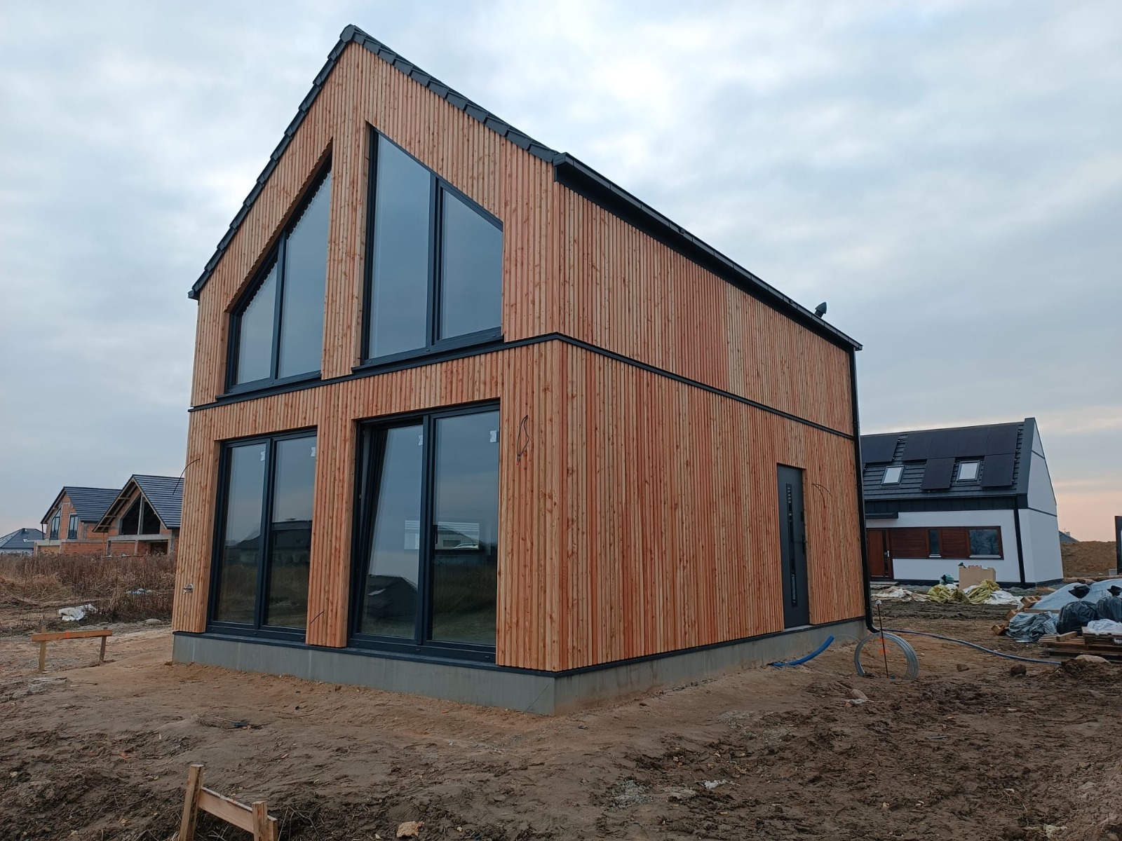 Nowoczesny dom z drewnianą elewacją i dużymi oknami, widoczny z zewnątrz na tle pochmurnego nieba. Charakterystyczne trójkątne okna na szczycie budynku.
