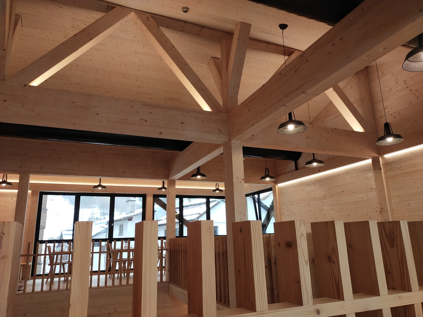 Wnętrze domu szkieletowego z drewnianą konstrukcją dachu i oświetleniem LED. Widok na okna z widokiem na ośnieżone budynki. Elementy wykończeniowe z jasnego drewna.