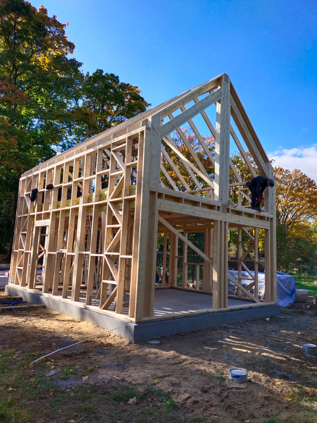 Konstrukcja szkieletowa domu w trakcie budowy, widoczne drewniane belki i dwóch pracowników na tle jesiennego krajobrazu i błękitnego nieba.