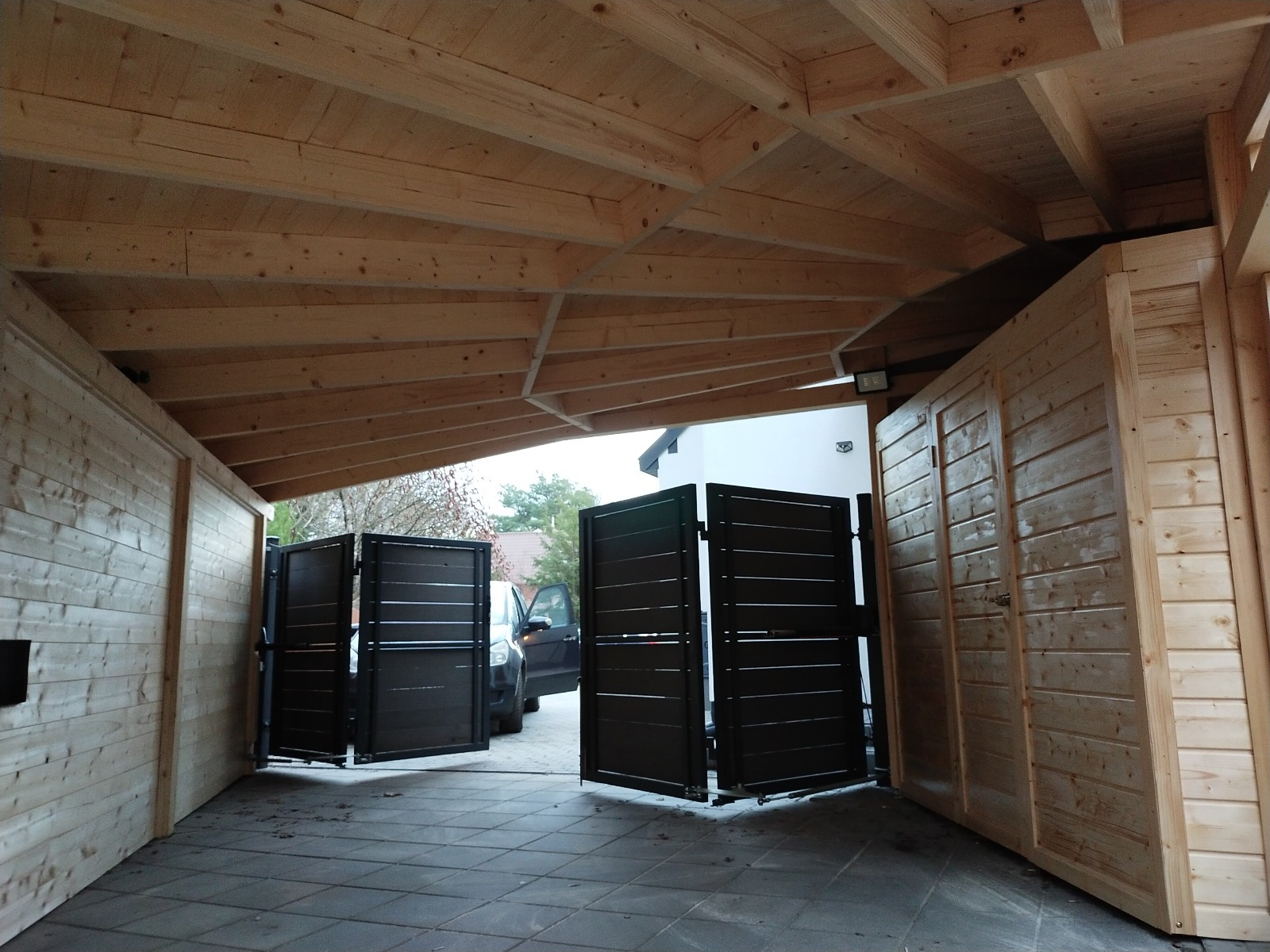 Drewniana wiata garażowa z otwieraną bramą wjazdową, widok od wewnątrz. Konstrukcja z jasnego drewna, brama w kolorze grafitowym. Na zewnątrz widoczny samochód.