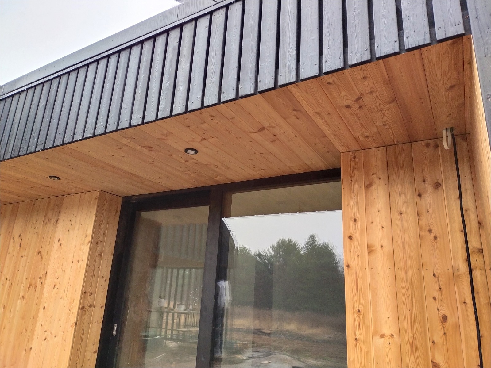 Nowoczesny dom szkieletowy z drewnianą elewacją i dużymi oknami, widok z dołu na fragment ściany i okapu z czarnymi elementami. Minimalistyczny design.