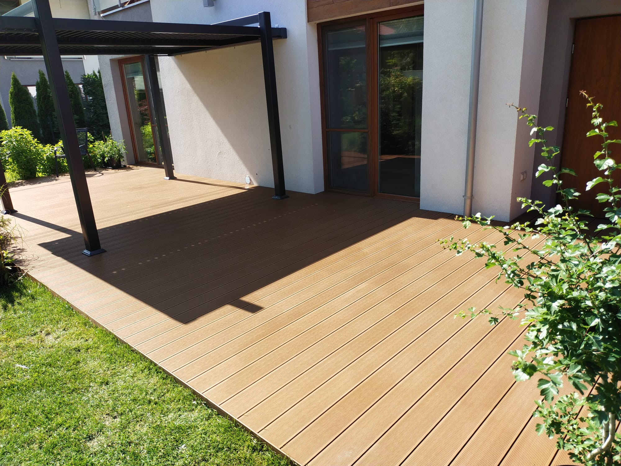 Nowoczesny taras z desek kompozytowych w odcieniach brązu, przylegający do jasnego budynku z ciemnymi oknami. Pergola rzuca cień na część tarasu, obok trawnik i zieleń.