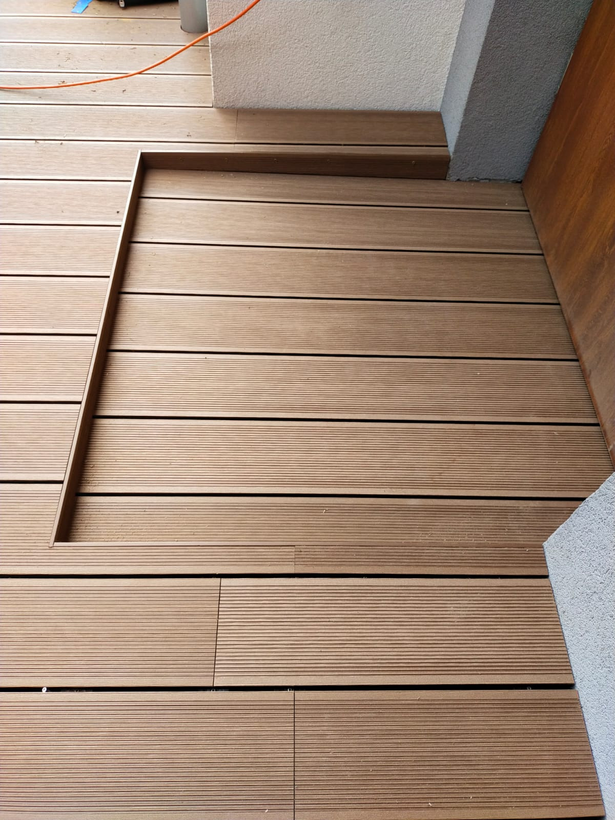 Beżowy taras z desek kompozytowych, z widocznym fragmentem ściany i drzwi. Kąt ujęcia podkreśla wzór desek i precyzję wykonania. Widoczne mocowania.