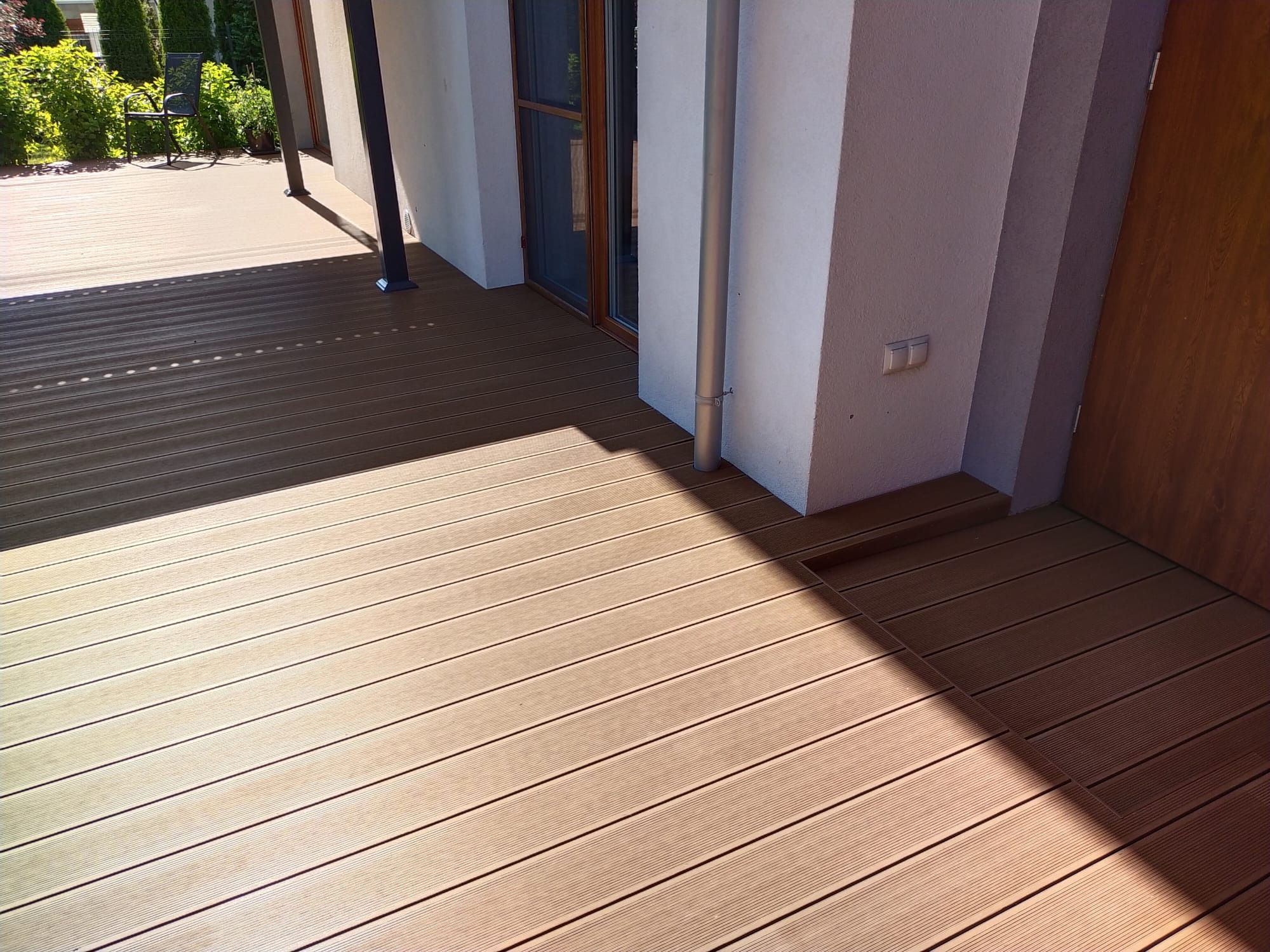 Jasnobrązowy taras z desek kompozytowych przylegający do jasnej elewacji budynku, z widoczną rynną i wejściem. Cień rzucany przez zadaszenie.