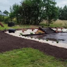 Projekt Wnętrz Sebastian Burski - Ukończony ogród z placem zabaw, pergolą i ścieżkami z białego żwiru. Widoczna świeżo przekopana ziemia pod trawnik. Całość w otoczeniu drzew i zieleni.