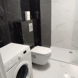 Projekt Wnętrz Sebastian Burski - Wykończona łazienka z czarno-białymi płytkami imitującymi marmur, podwieszana toaleta i pralka. Widoczne oświetlenie sufitowe i akcesoria łazienkowe.