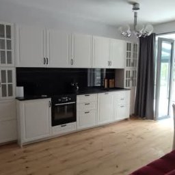 Projekt Wnętrz Sebastian Burski - Jasna kuchnia z białymi szafkami, czarnym blatem i drewnianą podłogą. Widoczne okno balkonowe z szarymi zasłonami i stół z krzesłami. Częściowo widoczna czerwona sofa.