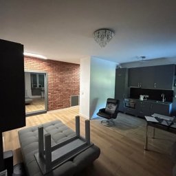 Projekt Wnętrz Sebastian Burski - Nowoczesne wnętrze mieszkania z ceglaną ścianą, szarą kuchnią i drewnianą podłogą. Widoczny fragment sofy i krzesło. Oświetlenie sufitowe w stylu glamour.