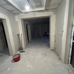 Projekt Wnętrz Sebastian Burski - Wnętrze w trakcie prac wykończeniowych: ściany z płyt karton-gips, surowy beton na podłodze, widoczne narzędzia i drabina w głębi. Okno z logo Veka.