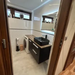 Nowoczesna łazienka z pralką obok szafki umywalkowej, biała mozaika ścienna i oświetlenie LED. Nietypowe połączenie funkcji prania i umywalni w jednym pomieszczeniu.