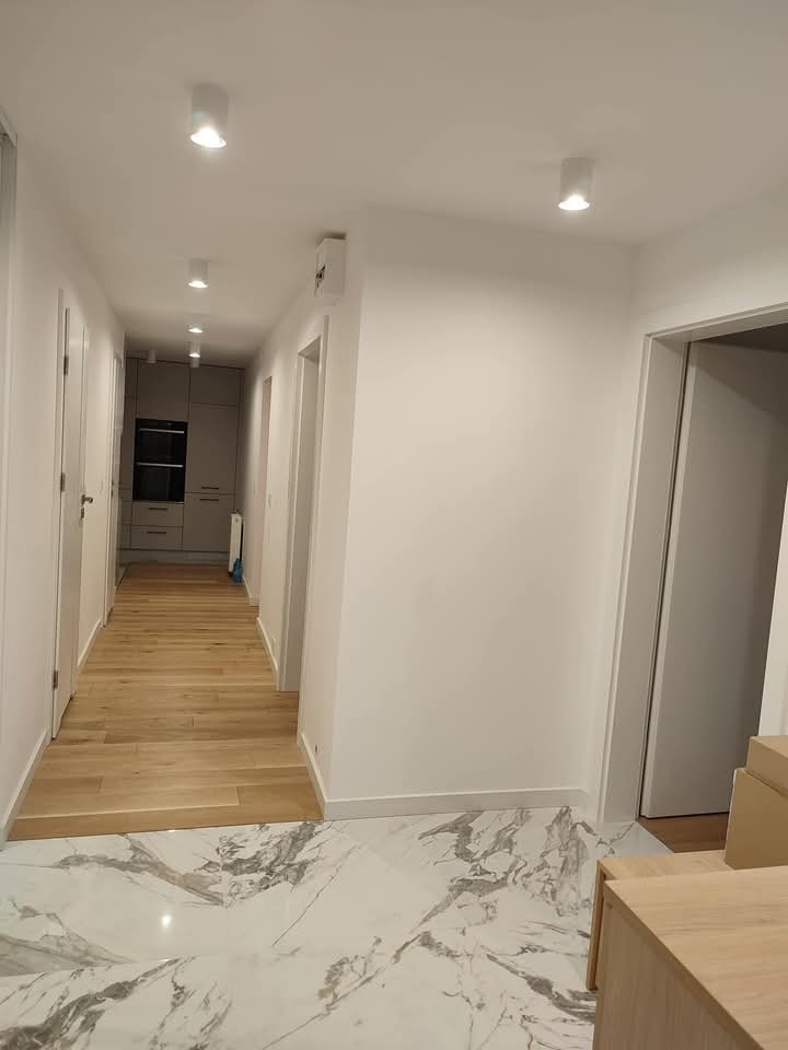 Wykończony korytarz z podłogą z marmuru i drewna. Widoczne drzwi, oświetlenie punktowe i fragment kuchni w tle. Ściany pomalowane na biało.