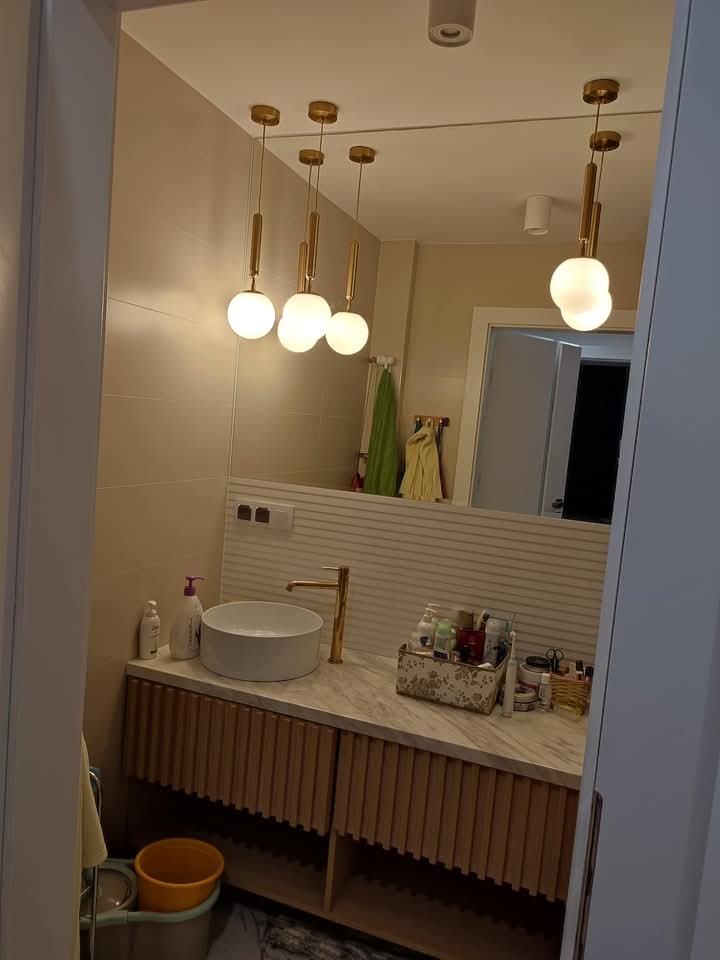 Nowoczesna łazienka z okrągłą umywalką, złotą armaturą i lampami kulami. Beżowe płytki na ścianach i szafka z lamelami. Lustro odbija fragment innego pomieszczenia.