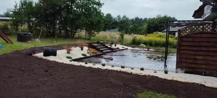 Ukończony taras z drewnianą podłogą, białym żwirem i schodami w ogrodzie. Widok na zieleń i drzewa w tle, z elementami architektury domu.