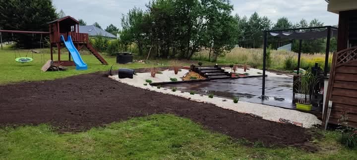 Ukończony ogród z placem zabaw, pergolą i ścieżkami z białego żwiru. Widoczna świeżo przekopana ziemia pod trawnik. Całość w otoczeniu drzew i zieleni.