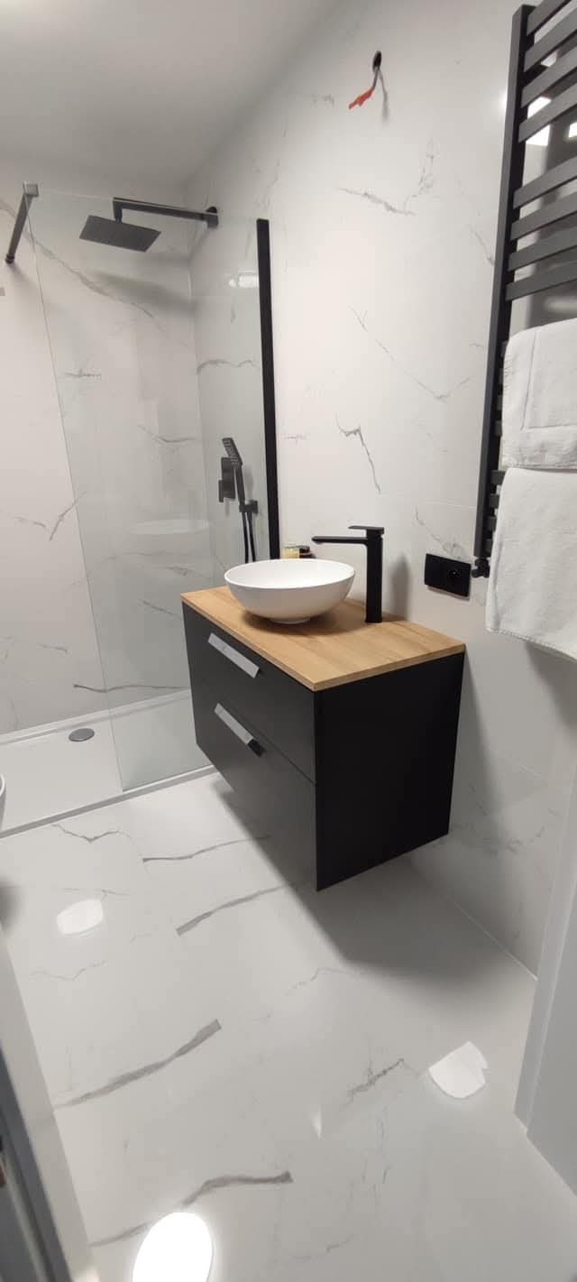 Nowoczesna łazienka z kabiną prysznicową typu walk-in, umywalką nablatową na szafce z drewnianym blatem i białymi płytkami imitującymi marmur.