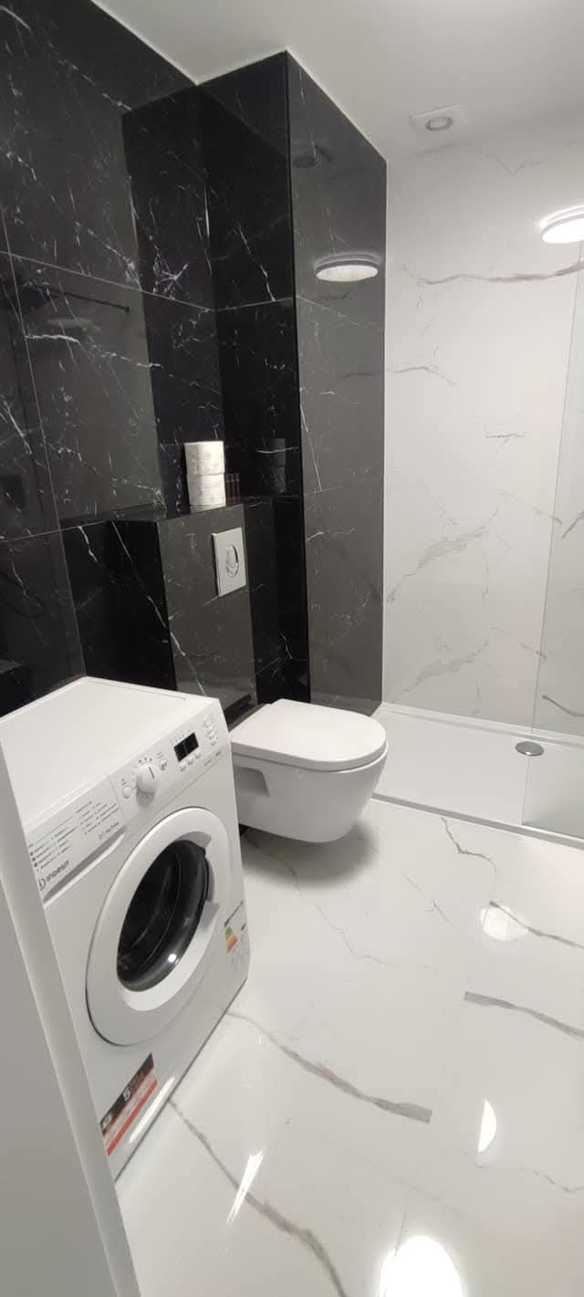 Wykończona łazienka z czarno-białymi płytkami imitującymi marmur, podwieszana toaleta i pralka. Widoczne oświetlenie sufitowe i akcesoria łazienkowe.