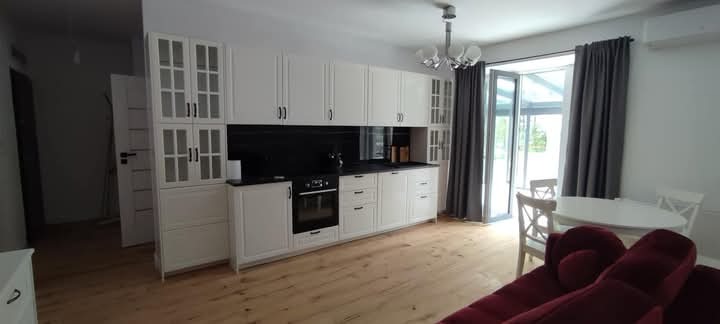 Jasna kuchnia z białymi szafkami, czarnym blatem i drewnianą podłogą. Widoczne okno balkonowe z szarymi zasłonami i stół z krzesłami. Częściowo widoczna czerwona sofa.