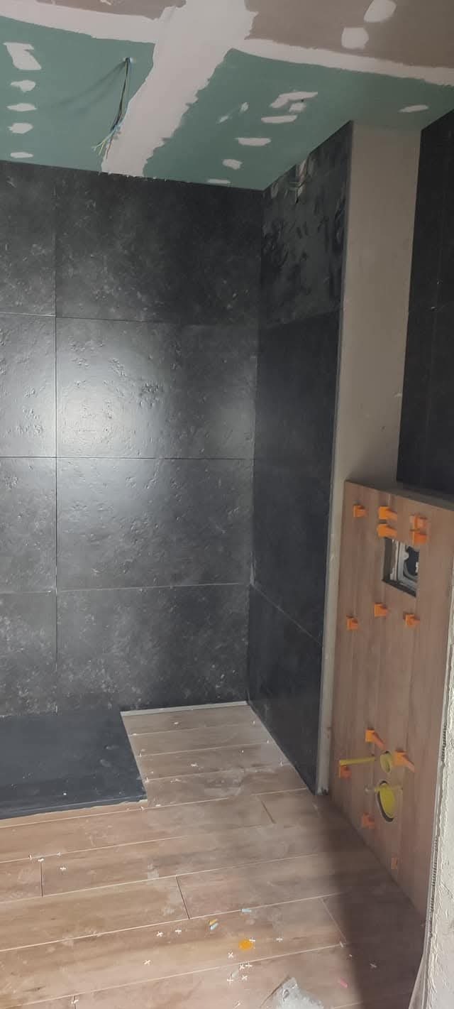 Ciemne płytki na ścianie w trakcie montażu w łazience, widoczne krzyżyki dystansowe. Podłoga częściowo wyłożona płytkami imitującymi drewno. Sufit z płyt karton-gips.