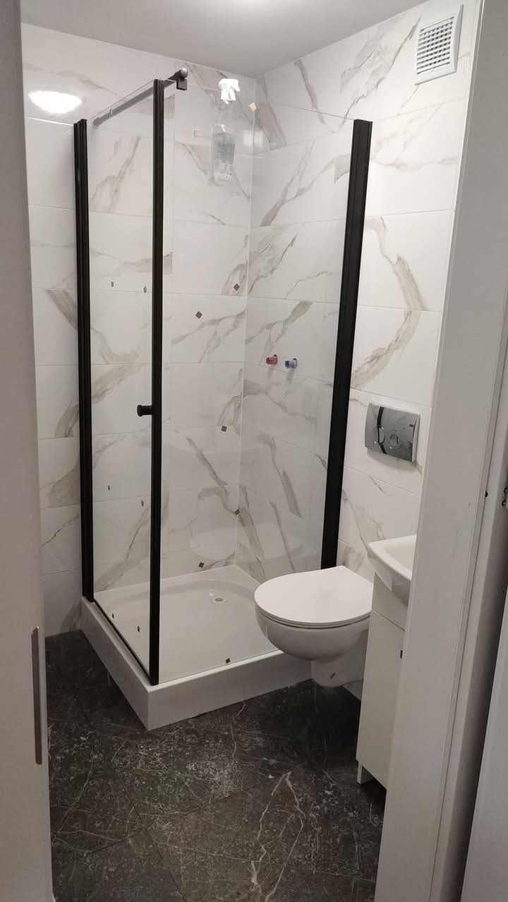 Nowoczesna łazienka z kabiną prysznicową typu walk-in, wykończona marmurowymi płytkami i czarnymi profilami. Widoczna toaleta i umywalka.