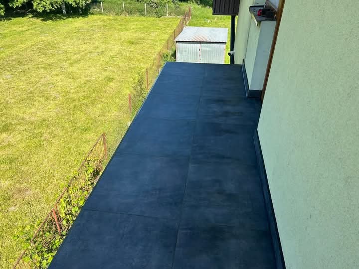 Wykończony balkon z ciemną, matową nawierzchnią z płytek, widok z góry, zieleń trawnika w tle, jasna elewacja budynku po prawej stronie.