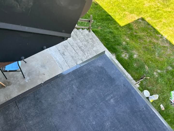 Widok z góry na fragment dachu pokrytego papą, z widocznym fragmentem trawnika i schodów. Prace dekarskie w toku, widoczne narzędzia i materiały budowlane.