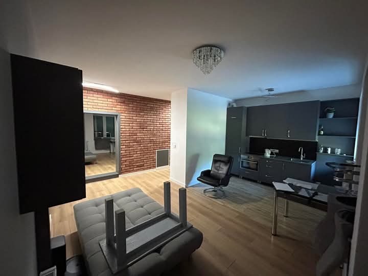 Nowoczesne wnętrze mieszkania z ceglaną ścianą, szarą kuchnią i drewnianą podłogą. Widoczny fragment sofy i krzesło. Oświetlenie sufitowe w stylu glamour.