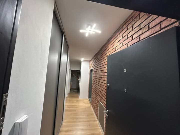 Korytarz z ceglaną ścianą i ciemnymi drzwiami wejściowymi z numerem '5'. Podłoga wyłożona panelami, sufit z nowoczesnym oświetleniem. Widoczne drzwi do pomieszczeń.
