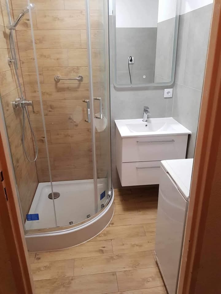 Jasna łazienka po remoncie: kabina prysznicowa z chromowanym zestawem, umywalka z szafką, lustro z podświetleniem i pralka. Drewnopodobne płytki na podłodze i ścianie.