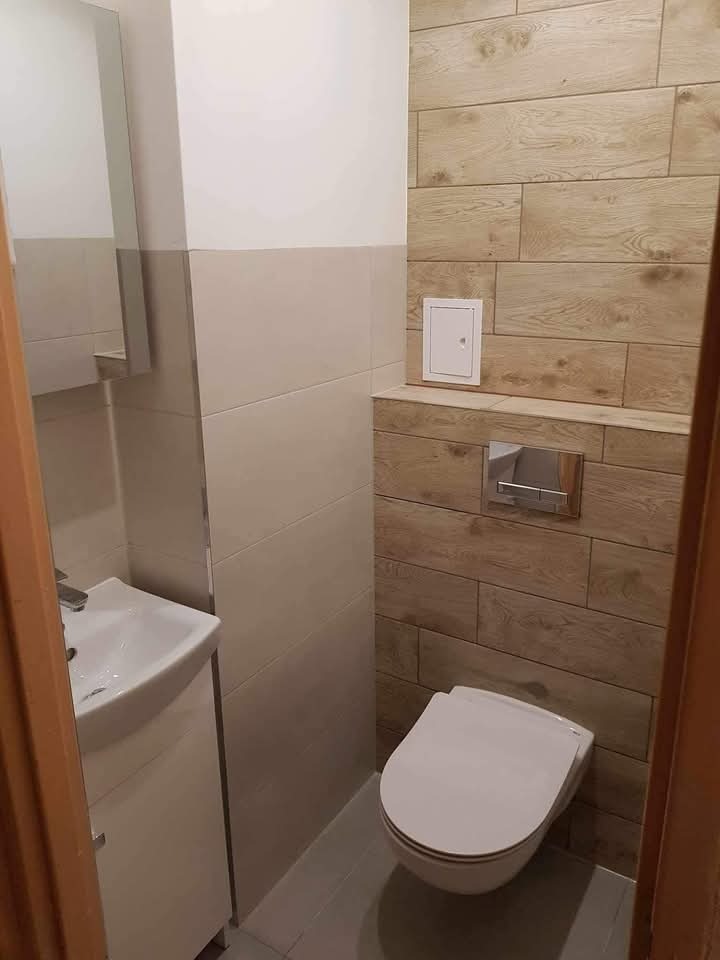 Nowoczesna toaleta z drewnopodobnymi płytkami, wiszącą miską WC i umywalką z szafką. Ściany wykończone w dwóch odcieniach płytek. Widoczny dostęp do instalacji.