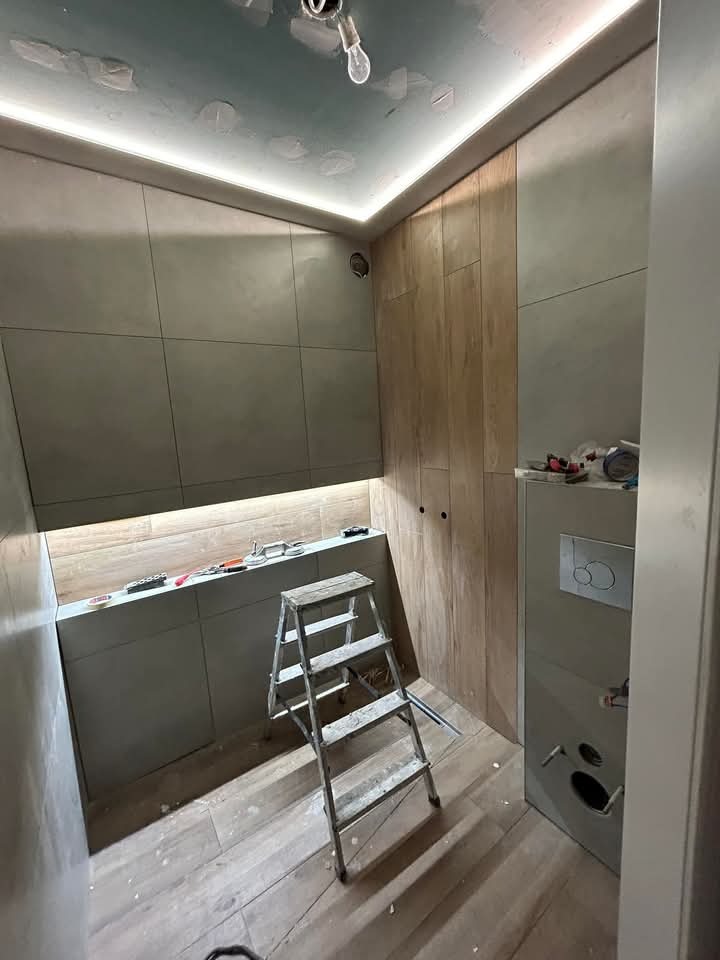 Remont łazienki w trakcie: płytki ścienne w odcieniach szarości i drewna, drabina, oświetlenie LED pod sufitem. Widoczne narzędzia i materiały wykończeniowe.