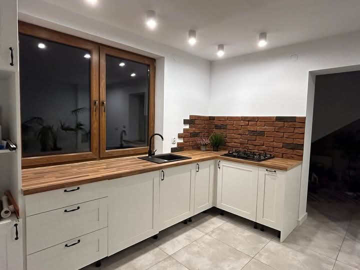 Jasna kuchnia z drewnianym blatem i ceglaną ścianą. Uwagę przykuwają duże okna z widokiem na ogród oraz minimalistyczne, czarne uchwyty szafek.