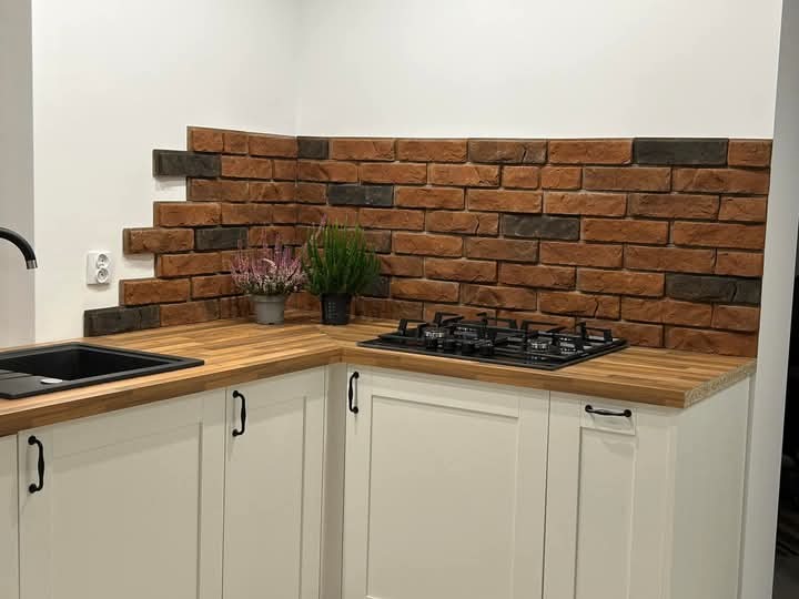 Kuchnia po remoncie: białe szafki z drewnianym blatem, ceglana ściana z efektem niedokończenia, czarny zlew i płyta gazowa. Rośliny w doniczkach dodają świeżości.