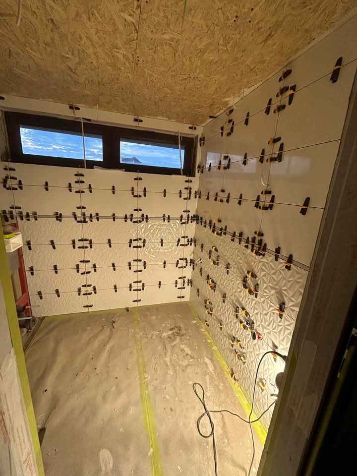 Wnętrze łazienki w trakcie układania białych, wzorzystych płytek. Widoczne krzyżyki dystansowe i sznurki poziomujące. Małe okno z widokiem na niebo i góry.