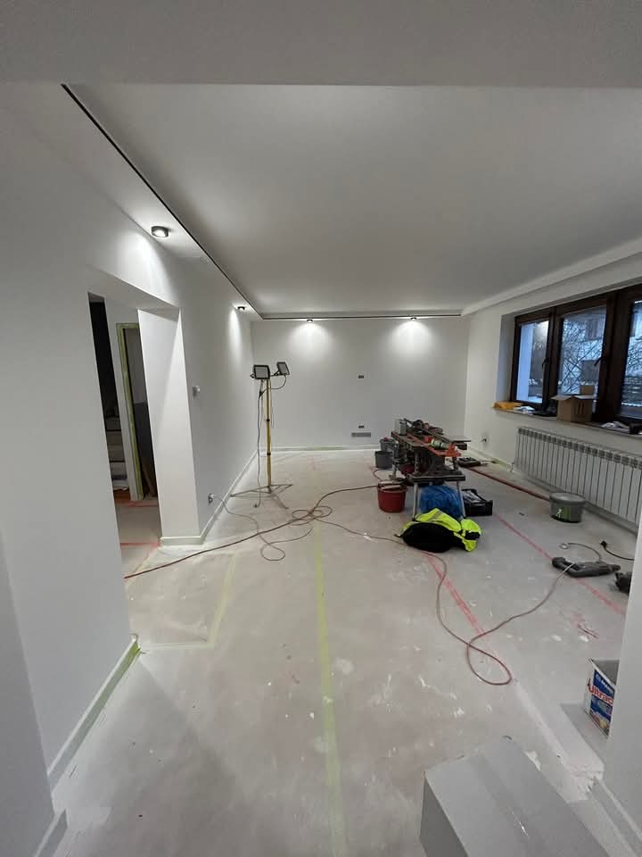 Białe wnętrze w trakcie remontu: widoczne narzędzia, lampy halogenowe, ślady po malowaniu i listwy sufitowe, podkreślające nowoczesny charakter przestrzeni.