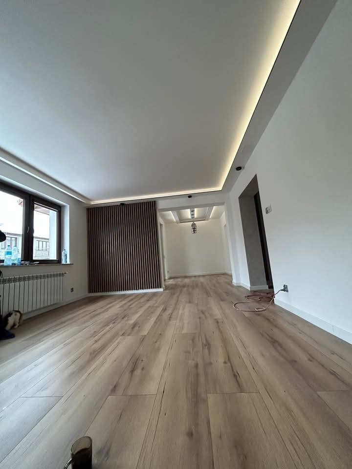 Jasne panele podłogowe w przestronnym wnętrzu z drewnianą ścianką działową i podwieszanym sufitem z oświetleniem LED. Widok na sąsiednie pomieszczenie.