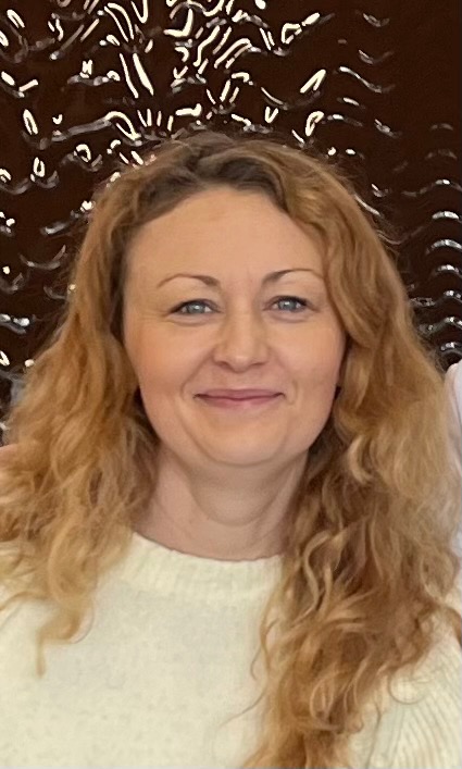 Portret uśmiechniętej kobiety o jasnych, kręconych włosach na tle dekoracyjnej ściany. Ubrana w jasny sweter, patrzy wprost w obiektyw.