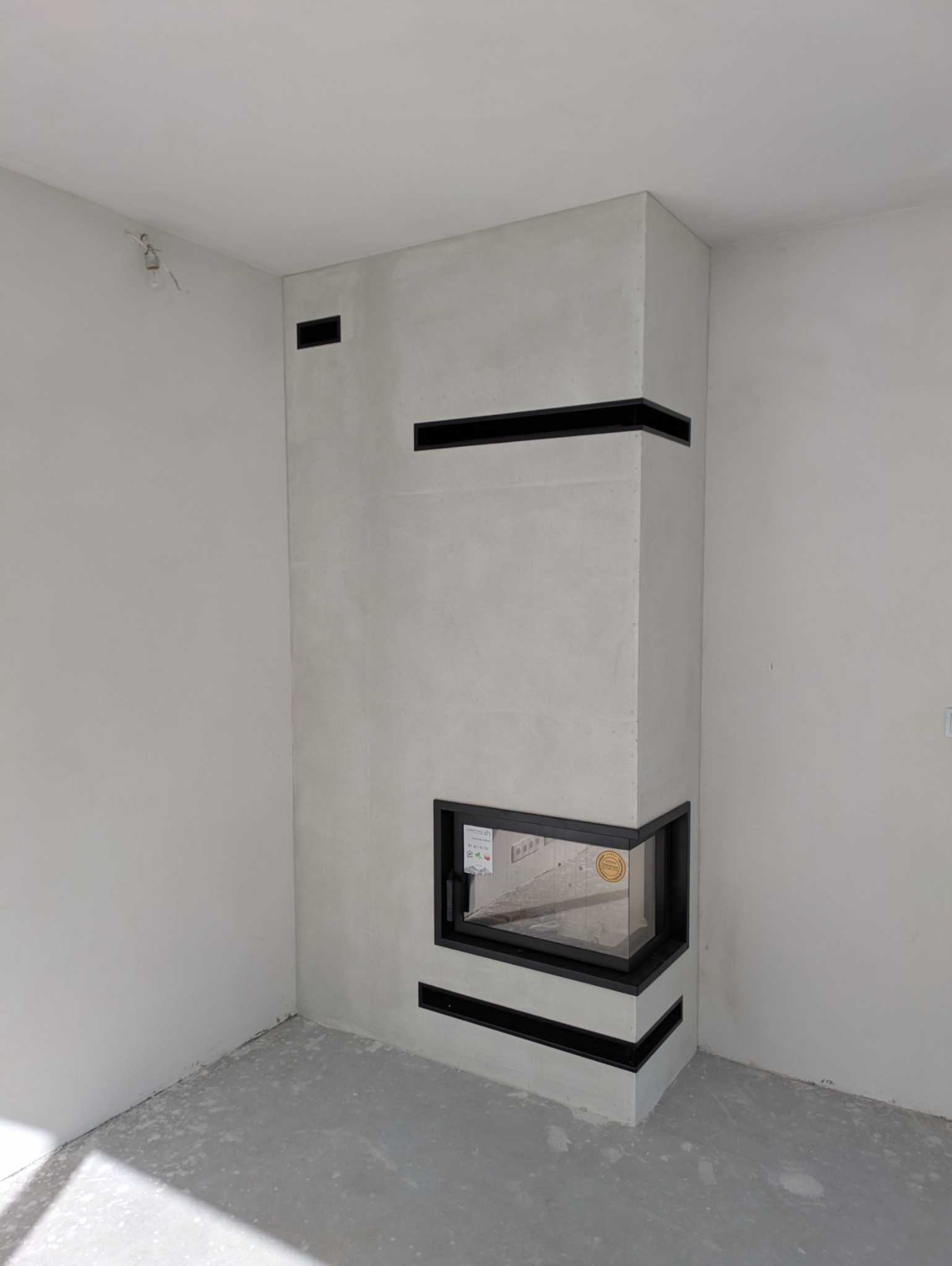 Narożny kominek z obudową w odcieniach szarości, z czarnymi, poziomymi akcentami, w surowym wnętrzu z widoczną instalacją elektryczną.