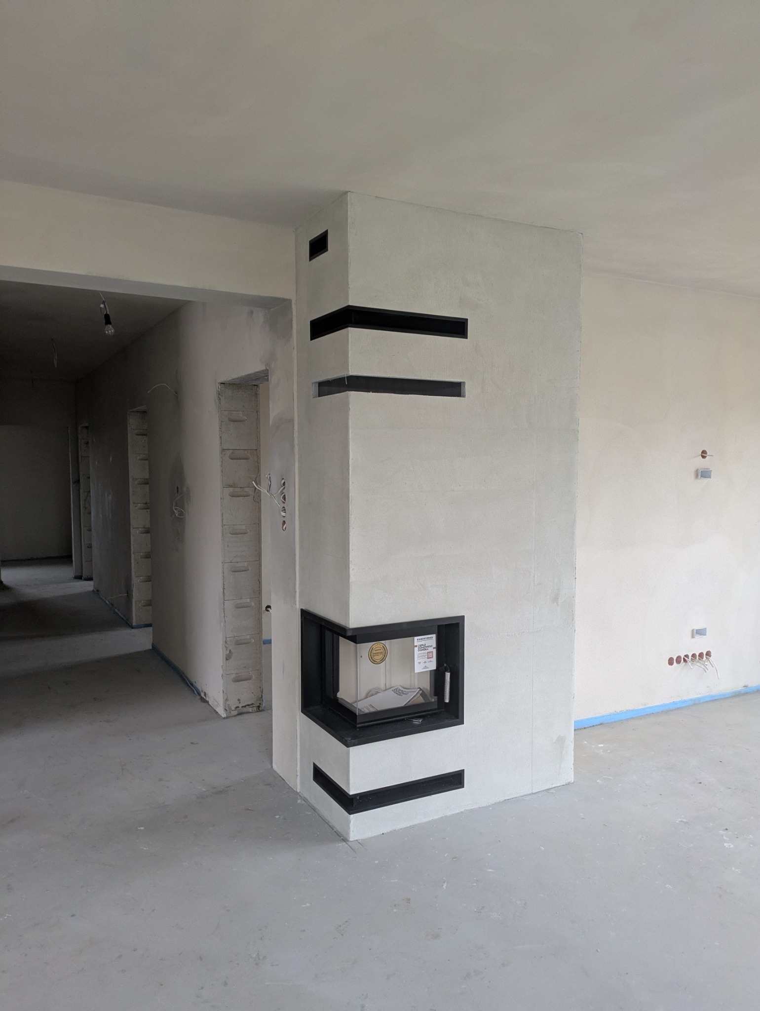 Narożny kominek z czarnymi elementami dekoracyjnymi w surowym wnętrzu, widok z perspektywy podłogi, w tle instalacje elektryczne i surowe ściany.