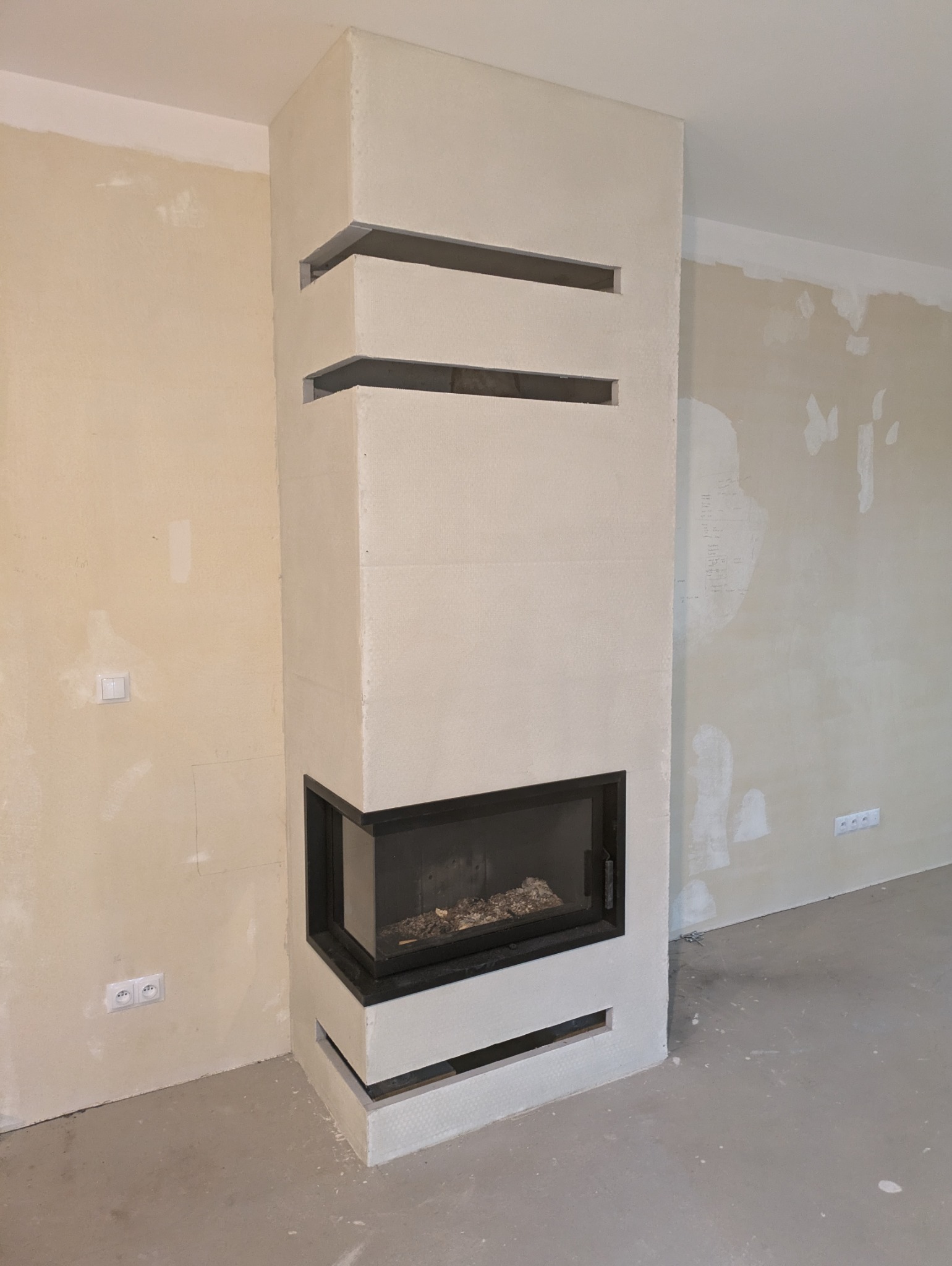 Nowoczesny, narożny kominek z wkładem i obudową z jasnego kamienia, z poziomymi, dekoracyjnymi szczelinami wentylacyjnymi. Wnętrze w trakcie wykończenia.