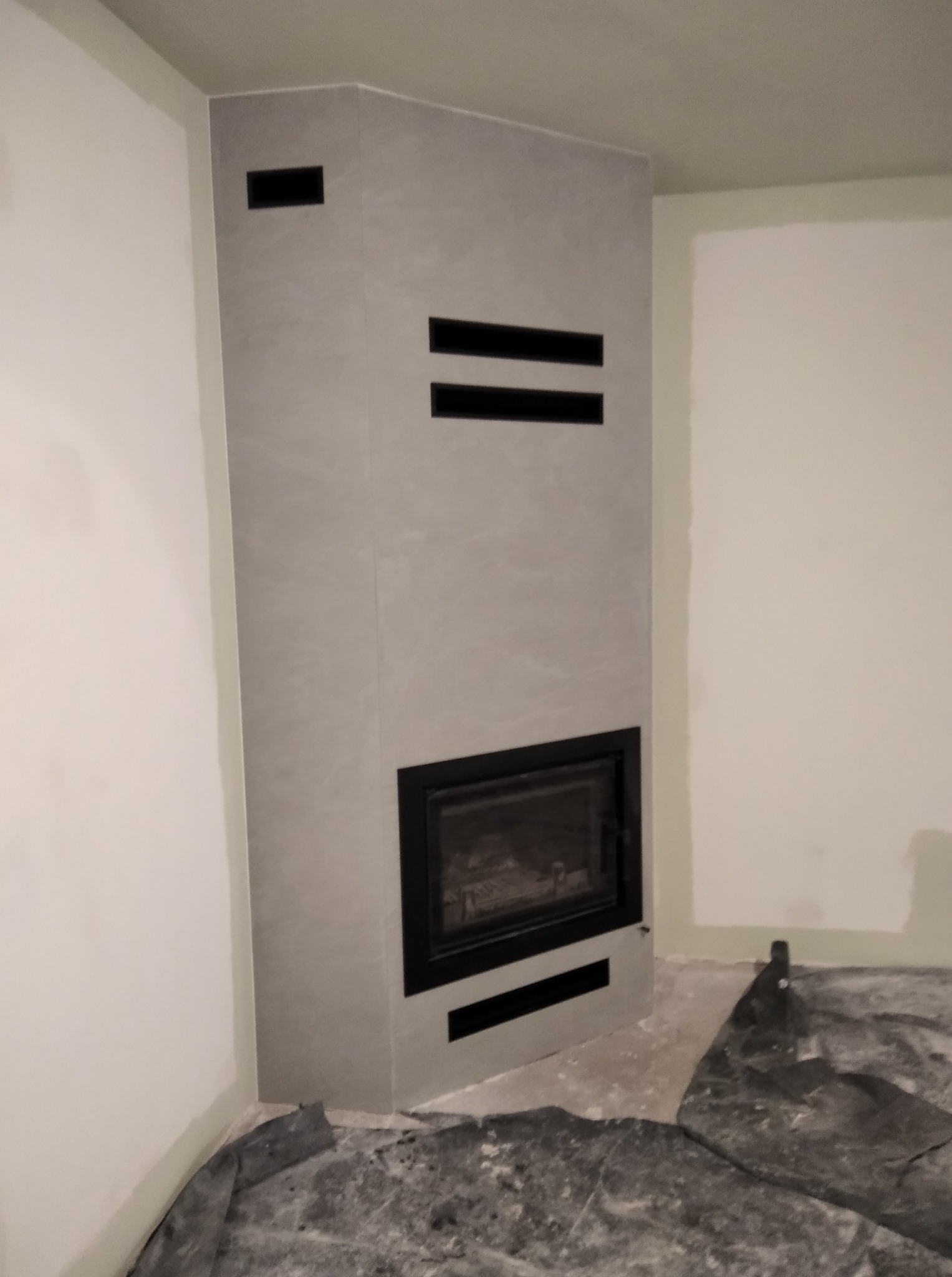 Nowoczesny, szary kominek narożny z czarnym wkładem i poziomymi wlotami powietrza w surowym wnętrzu, zabezpieczony folią malarską na podłodze.