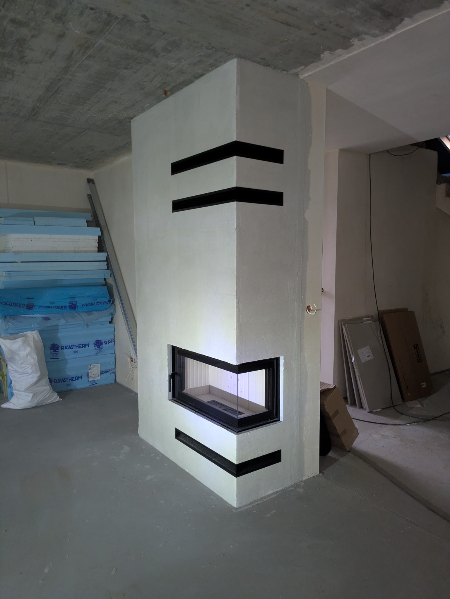 Nowoczesny, narożny wkład kominkowy z czarnymi akcentami w surowym wnętrzu. Widoczne materiały budowlane, betonowy strop i instalacja elektryczna.