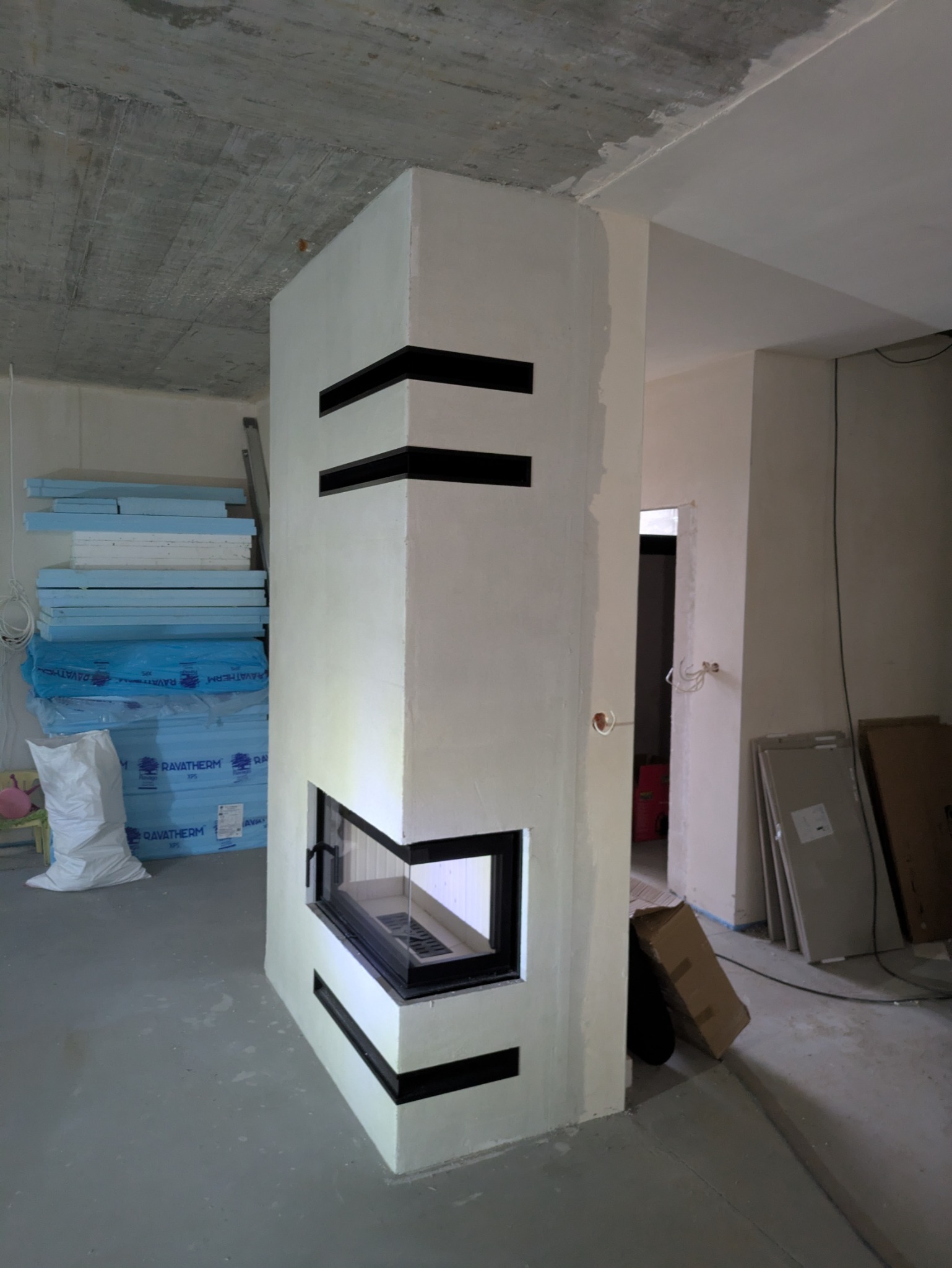 Nowoczesny, narożny kominek z czarnymi, poziomymi wstawkami w surowym wnętrzu. Widoczne materiały budowlane i betonowy strop.